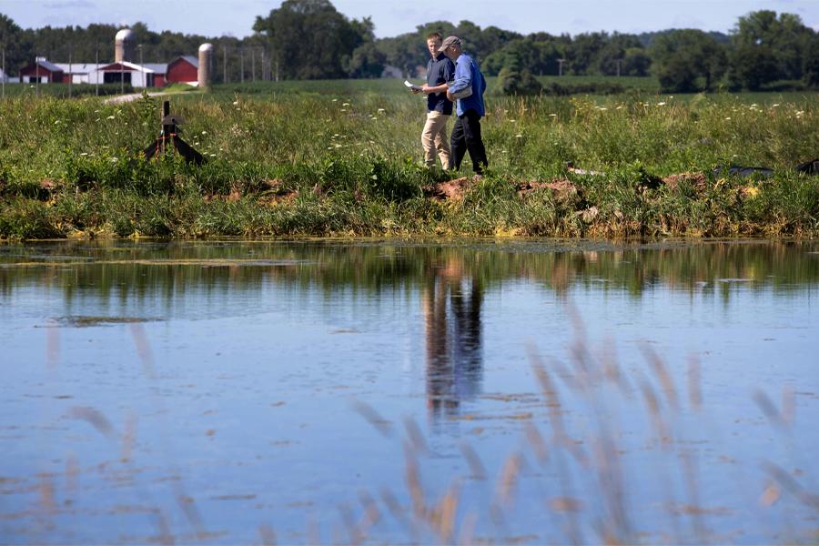 Two people walk by a waterway with a farm in the background.